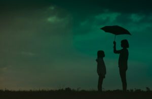 A woman with an umbrella shields a young person from the rain atop a grassy hill
